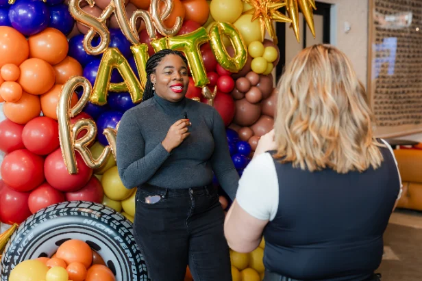 A woman speaking to camera against a balloon backdrop while Natalie directs