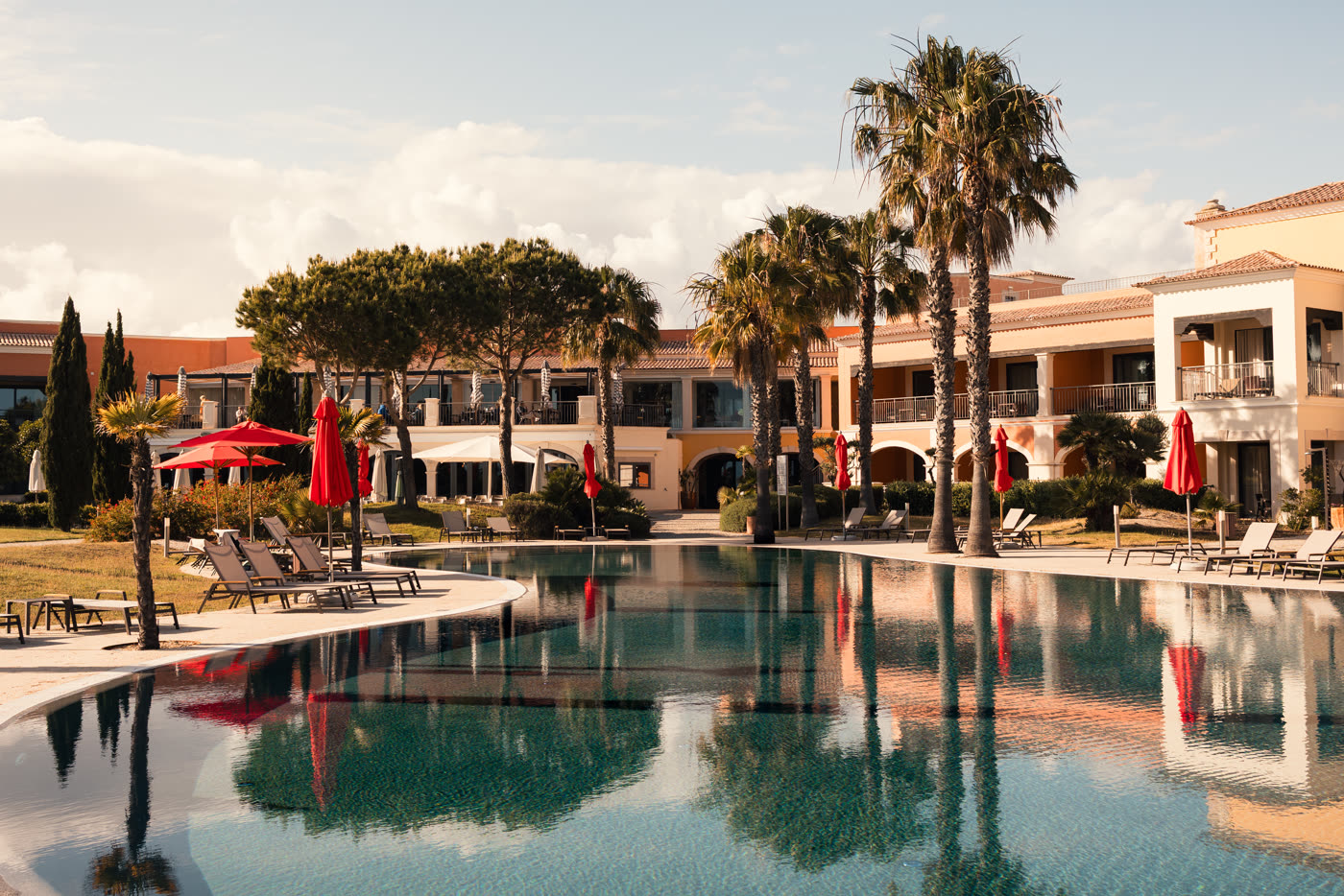 Resort pool at golden hour, palms and red parasols reflected in the water
