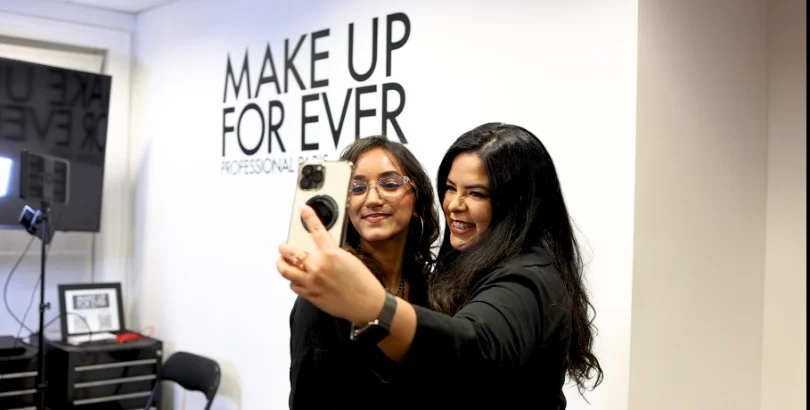 Two people laughing while filming a phone selfie inside a beauty brand's studio space