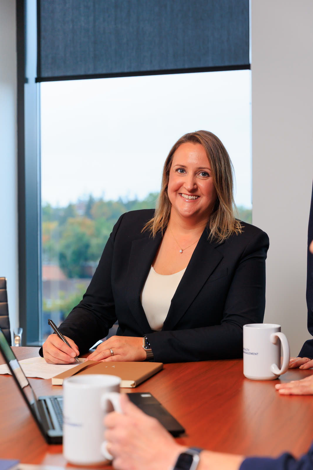 Woman in a navy blazer smiling at a boardroom table, pen in hand